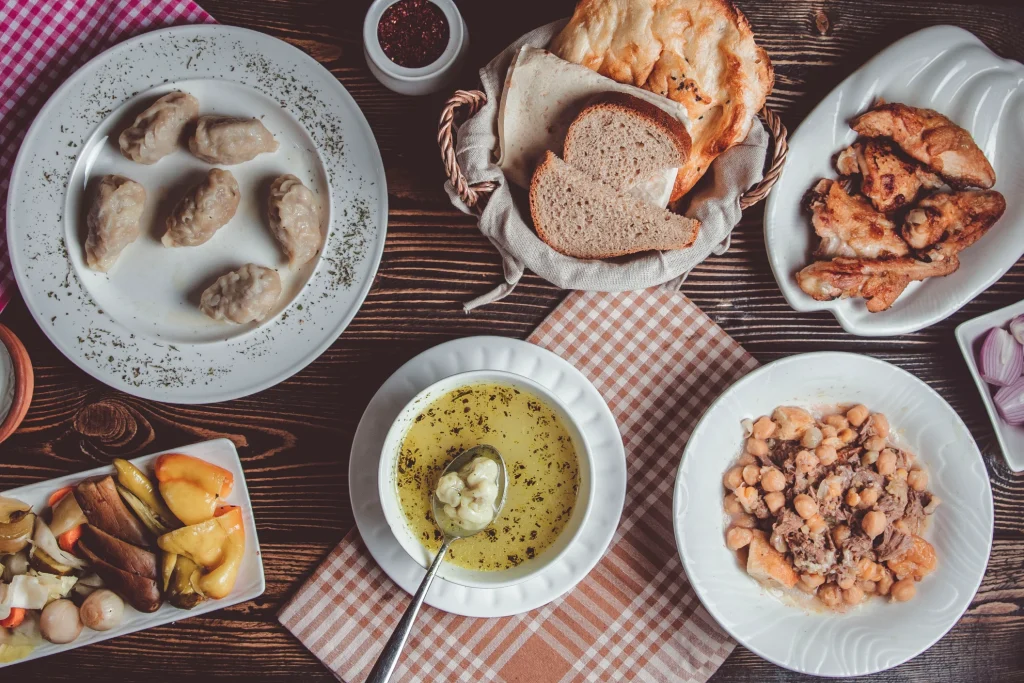 Vista superior de uma mesa rústica de madeira escura com pratos da culinária tradicional. Há uma sopa caseira com ervas sobre um pano xadrez, um prato com guisado de grão-de-bico e carne, uma travessa de frango assado, uma cesta de vime com fatias de pão regional, rissóis ou pastéis cozidos e acompanhamentos de picles.