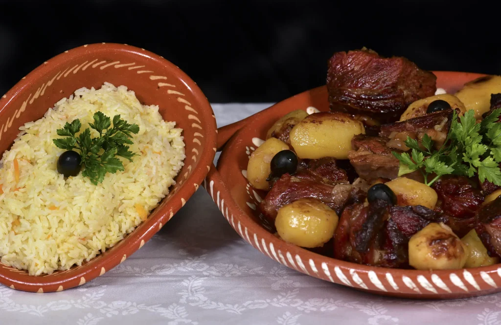 Dois pratos de barro: um com arroz amarelo decorado com salsa e azeitona, e outro com carne assada, batatas pequenas e azeitonas.