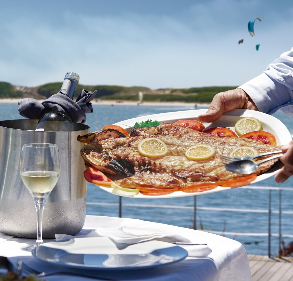 Prato de peixe grelhado com limão e tomate, colocado numa mesa ao ar livre com um copo de vinho branco e vista para o rio ao fundo.