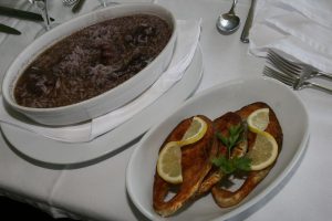 Taça com arroz de sável em caldo escuro ao lado de prato com duas postas de peixe frito decoradas com limão e salsa.