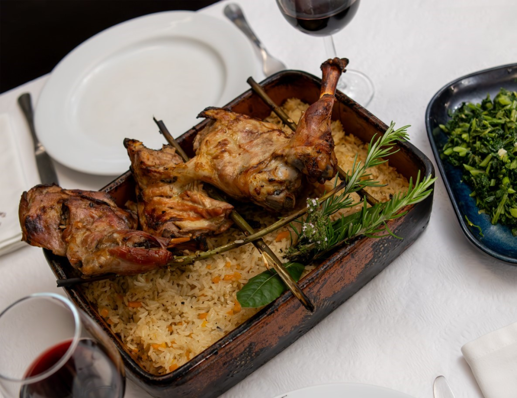 Travessa com carne assada colocada sobre arroz com ervas, acompanhada por um prato de legumes verdes e dois copos de vinho tinto.