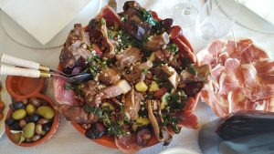 Travessa com Cozido à Portuguesa, incluindo carnes, enchidos, batatas, couves e feijão, ao lado de presunto fatiado e azeitonas.