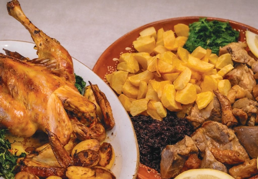 Prato com galo assado e batatas ao lado de outro prato com rojões, batata frita em cubos, carne e legumes verdes.