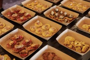Tabuleiros de papel com pequenas porções de petiscos variados, incluindo bases de pão, carnes e fritos.