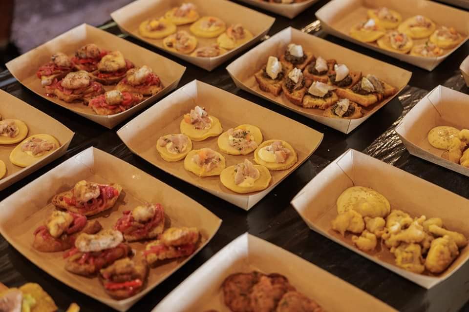 Tabuleiros de papel com pequenas porções de petiscos variados, incluindo bases de pão, carnes e fritos.