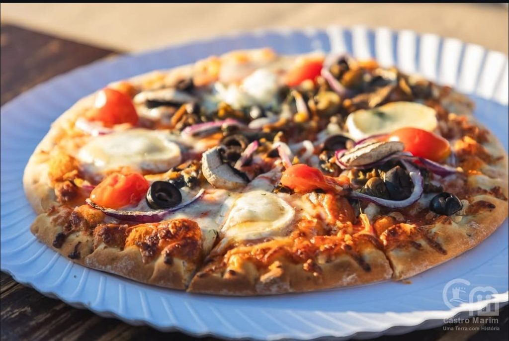 Pizza com queijo, cogumelos, azeitonas, tomate e cebola roxa num prato branco, com logótipo de Castro Marim no canto.
