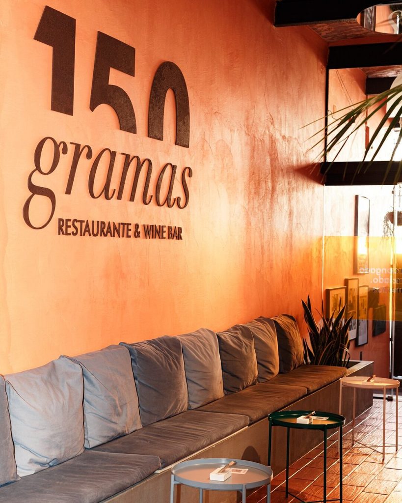 Interior do restaurante com parede em tons quentes, logótipo “150 Gramas Restaurante & Wine Bar” e mesas com bancos almofadados.