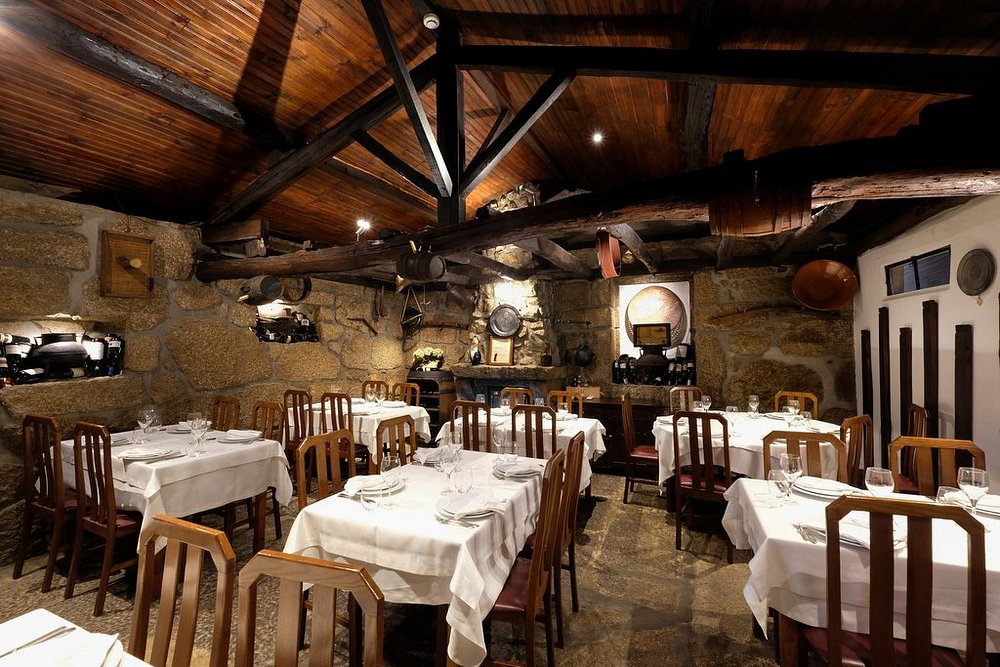 Sala de restaurante com mesas cobertas por toalhas brancas, paredes em pedra e teto com vigas de madeira.