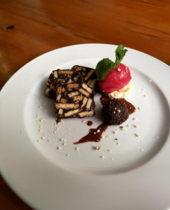 Prato com duas fatias de bolo de bolacha, uma bola de gelado vermelho com hortelã e uma trufa de chocolate.