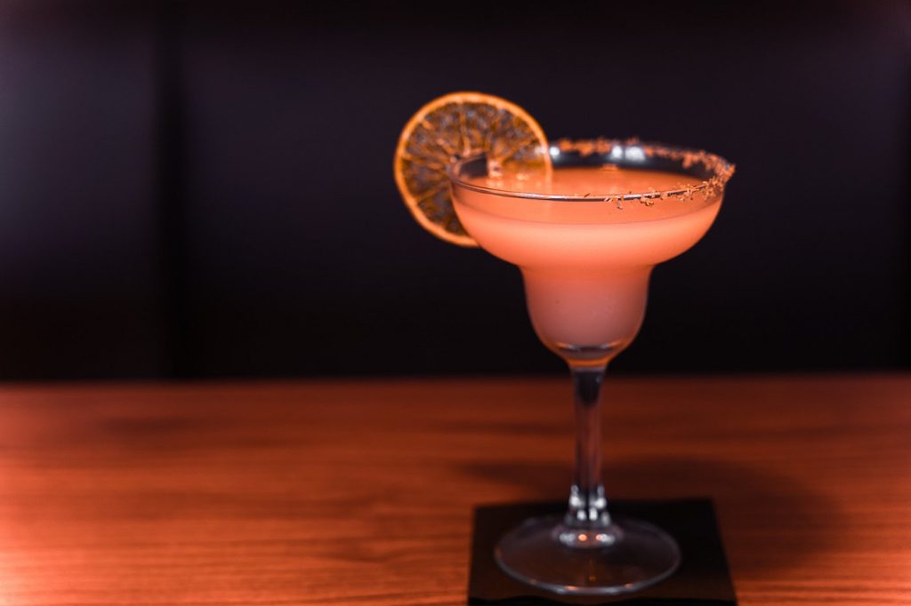 Cocktail rosa com borda açucarada e rodela de citrino desidratado num ambiente escuro.
