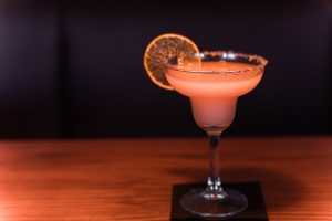 Cocktail rosa com borda açucarada e rodela de citrino desidratado num ambiente escuro.