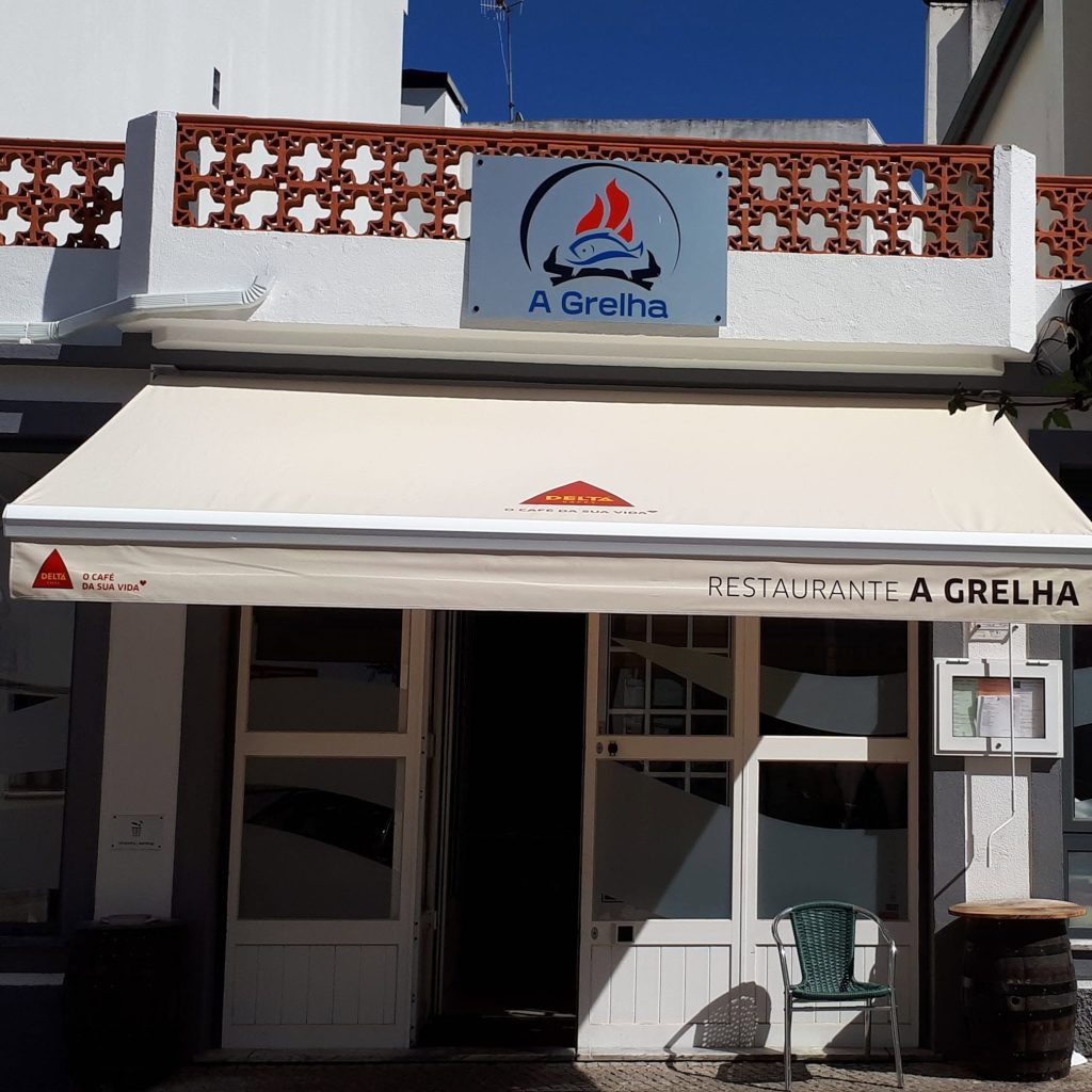 A imagem mostra a fachada do Restaurante A Grelha, em Arganil. O edifício apresenta paredes claras, um toldo com a marca Delta e o nome do restaurante, além do logótipo com um peixe estilizado. Em frente à entrada existe uma mesa em formato de barril e uma cadeira verde.
