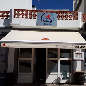 A imagem mostra a fachada do Restaurante A Grelha, em Arganil. O edifício apresenta paredes claras, um toldo com a marca Delta e o nome do restaurante, além do logótipo com um peixe estilizado. Em frente à entrada existe uma mesa em formato de barril e uma cadeira verde.