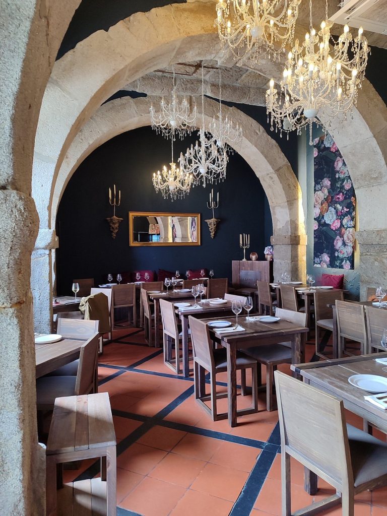 Sala de restaurante com arcos em pedra, mesas de madeira, lustres de cristal e decoração clássica.