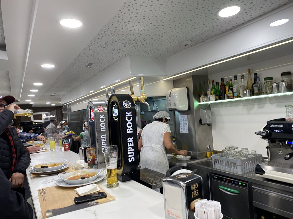 Balcão de restaurante com clientes sentados e em pé, com torneiras de cerveja, pratos, copos e funcionário ao fundo.