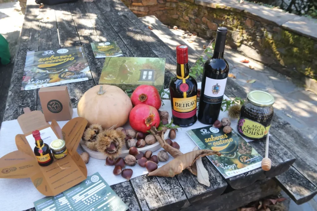 Mesa rústica exibe abóbora, romãs, castanhas, mel, vinho, licor Beirão e folhetos do Festival Sabores de Outono, em ambiente ao ar livre;