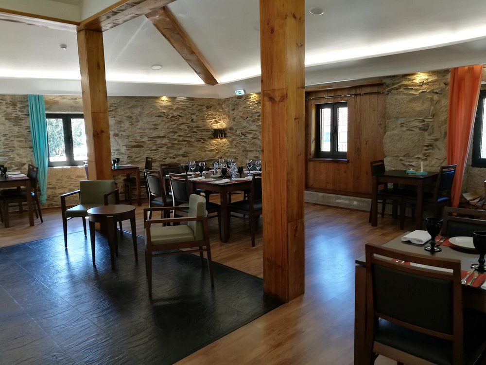 Sala com paredes de pedra, mesas montadas com pratos e copos, cortinas coloridas e teto com vigas de madeira.