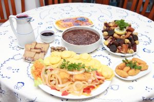 Mesa com toalha branca bordada e vários pratos tradicionais, incluindo carnes, fritos, doces e bebida escura em jarro.