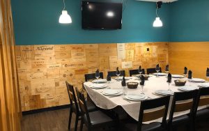 Sala com mesa retangular posta, cadeiras pretas e paredes decoradas com painéis de marcas de vinho.