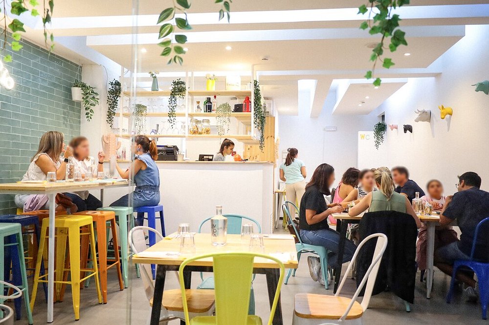 Interior do Cais do Rio com cadeiras coloridas, mesas ocupadas por clientes e elementos decorativos como plantas e prateleiras.