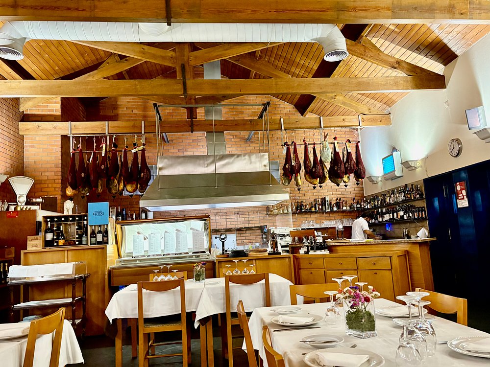 Sala de restaurante com mesas postas, presuntos pendurados no teto e balcão com garrafas expostas.