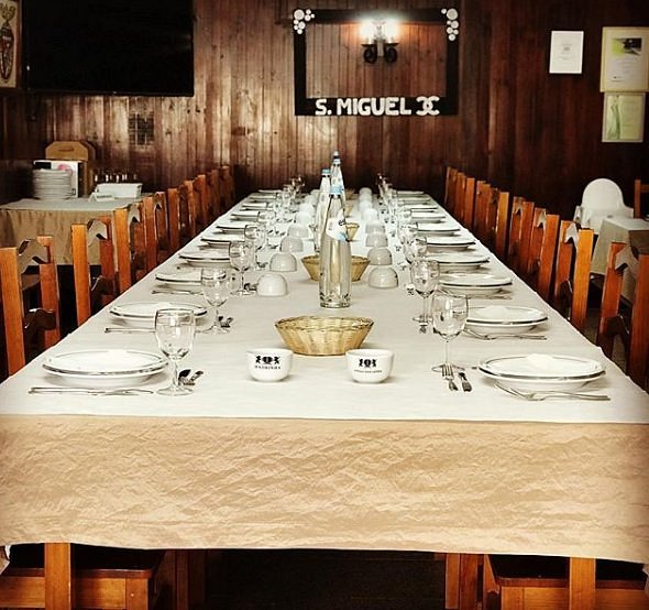 Mesa comprida com pratos, taças, copos e talheres, preparada para um grupo numeroso.