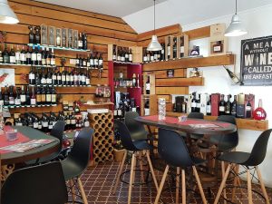 Sala com prateleiras de vinhos, mesas redondas, cadeiras pretas e iluminação pendente.