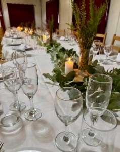 Mesa comprida preparada para evento, com toalha branca, folhagem verde ao centro, velas acesas e copos alinhados.