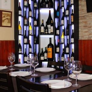 Mesa com pratos, guardanapos e copos de vinho, com garrafeira iluminada cheia de garrafas ao fundo.