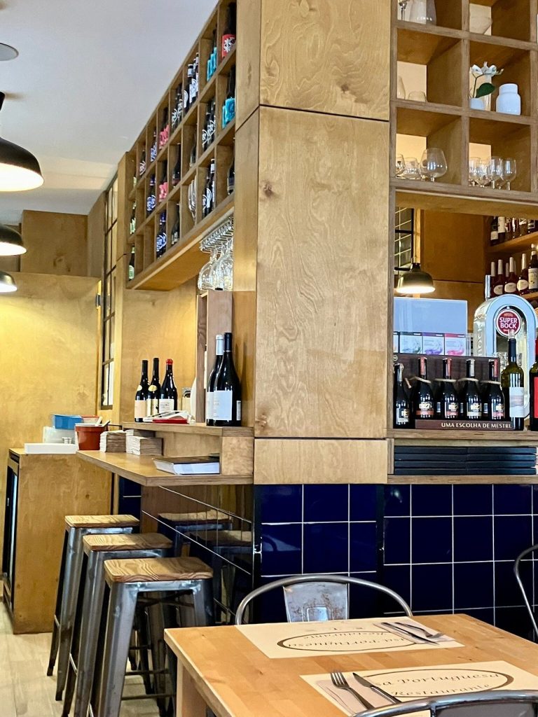 Sala com prateleiras de bebidas, balcão de madeira, mesas com individuais e iluminação pendente.