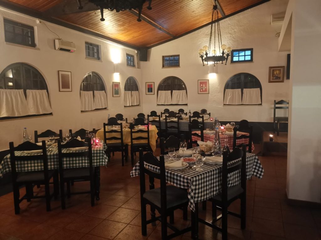 Sala de refeições com mesas de toalhas coloridas, cadeiras escuras e janelas arqueadas num ambiente acolhedor.