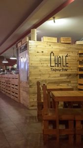 A imagem mostra o interior do Chalé Tapas Bar, com paredes revestidas em madeira clara e mesas de madeira distribuídas pela sala. Ao fundo vê‑se o balcão com iluminação suspensa e uma pessoa a trabalhar. Na parede está o nome “Chalé Tapas Bar” em letras pretas. O ambiente é acolhedor, com decoração simples e rústica.