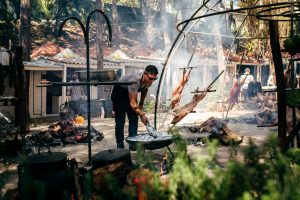 Assados inteiros suspensos e panelas de ferro ao lado de fogueira, com cozinheiro a preparar alimentos.