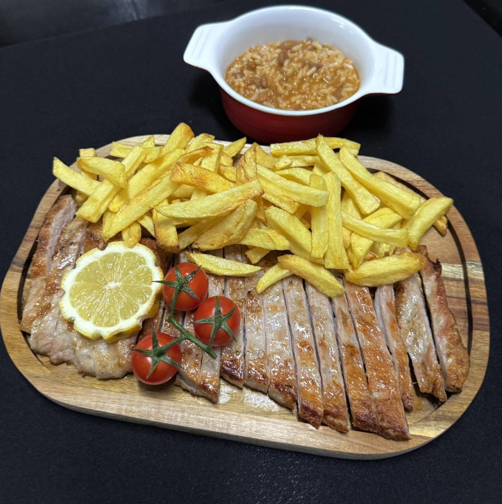 Tábua com carne grelhada, batatas fritas, limão e tomates, acompanhada por taça de arroz com molho.