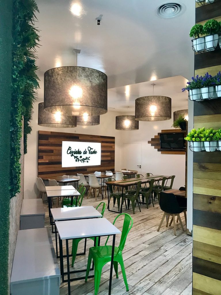 Sala de restaurante com mesas de madeira, cadeiras verdes e brancas, iluminação suspensa e parede com logótipo Cozinha do Tacho.