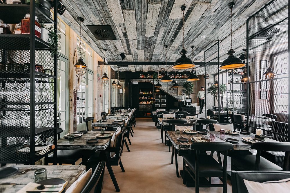 Sala de restaurante com mesas de madeira, cadeiras pretas, iluminação suspensa e divisórias metálicas.