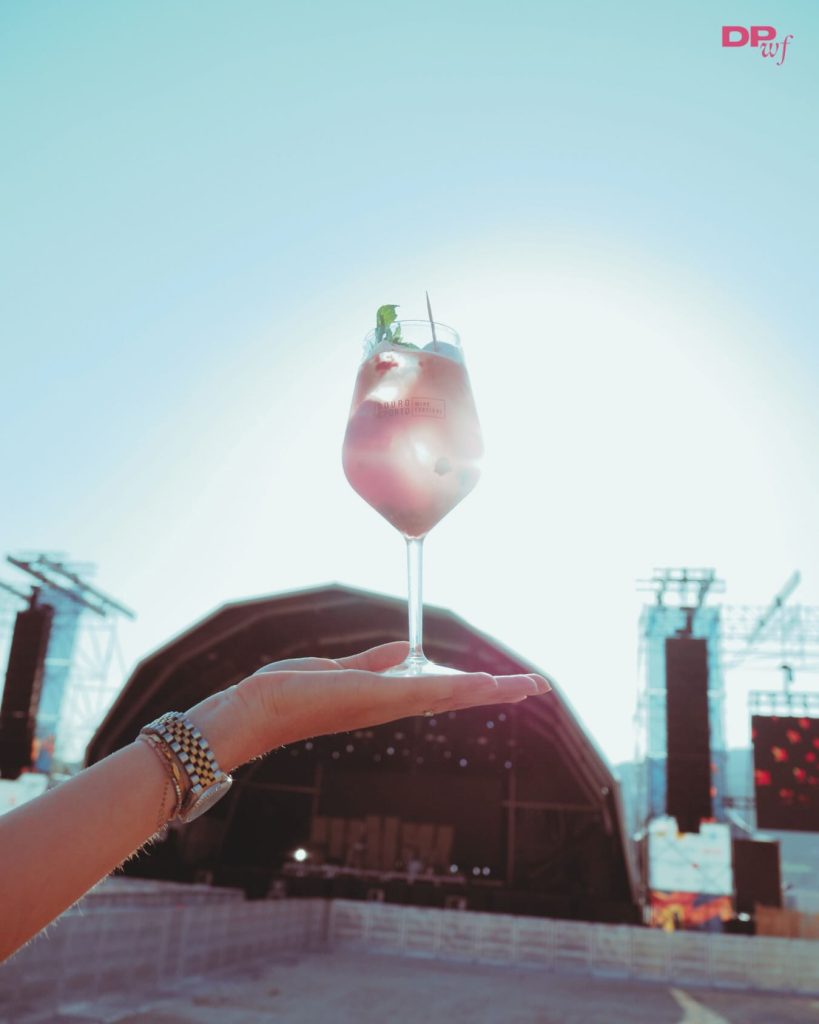 Mão a segurar copo com bebida rosa, gelo e hortelã, com palco de concerto ao fundo.
