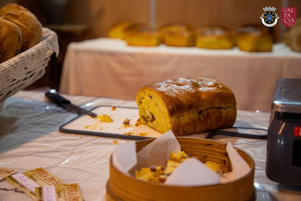 Pão rústico com frutos secos fatiado numa tábua, com cesto e pães inteiros ao fundo.
