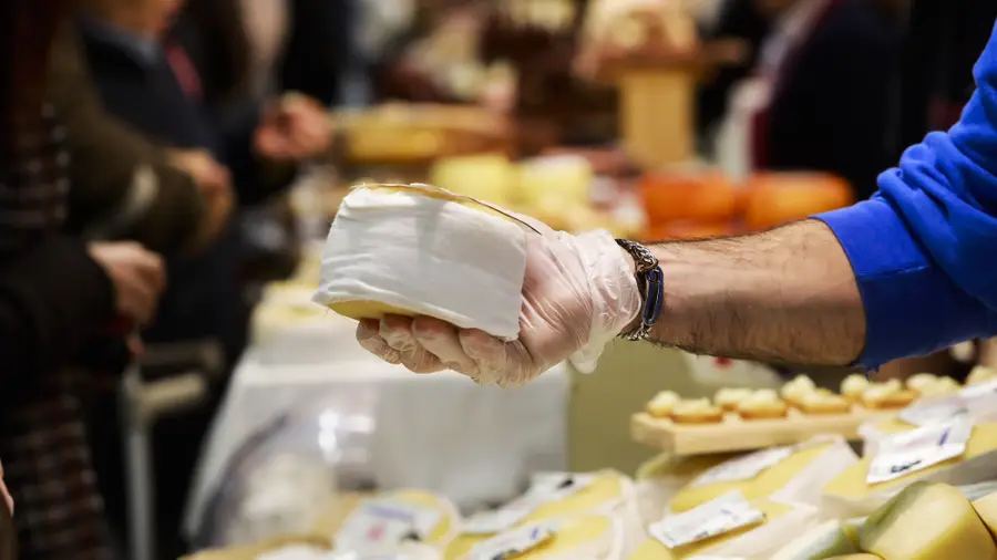 Mão com luva a segurar um pedaço de queijo embrulhado em papel, com vários queijos expostos numa banca.