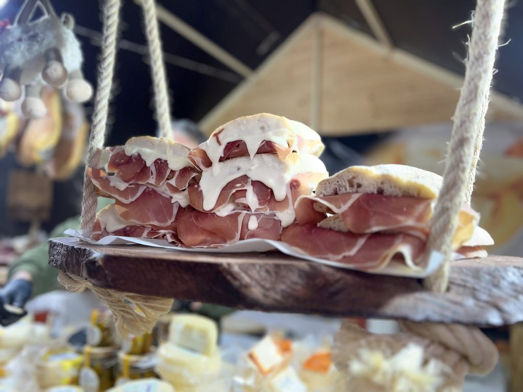 Sandes com pão rústico, presunto e queijo cremoso empilhadas em tabuleiro de madeira suspenso por cordas.