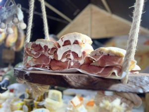 Sandes com pão rústico, presunto e queijo cremoso empilhadas em tabuleiro de madeira suspenso por cordas.