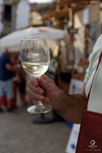 Pessoa a segurar copo de vinho branco com logótipo “Provesende aldeia vinhateira” num mercado ao ar livre.