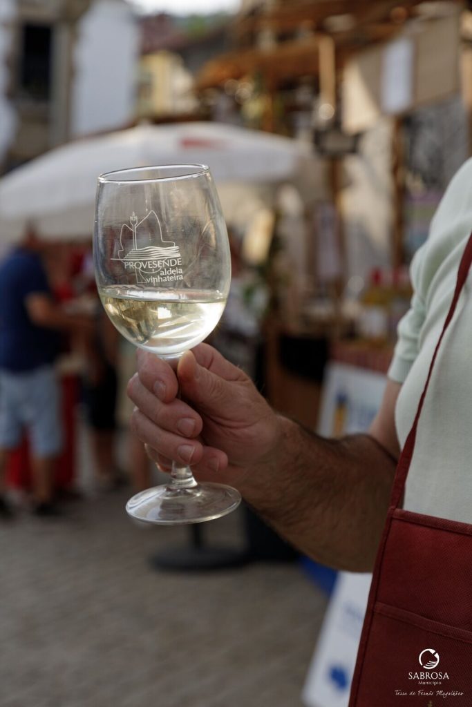 Pessoa a segurar copo de vinho branco com logótipo “Provesende aldeia vinhateira” num mercado ao ar livre.