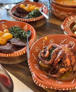Travessa de barro com polvo assado e batatas a murro ao lado de pratos tradicionais de barro com carne e migas.