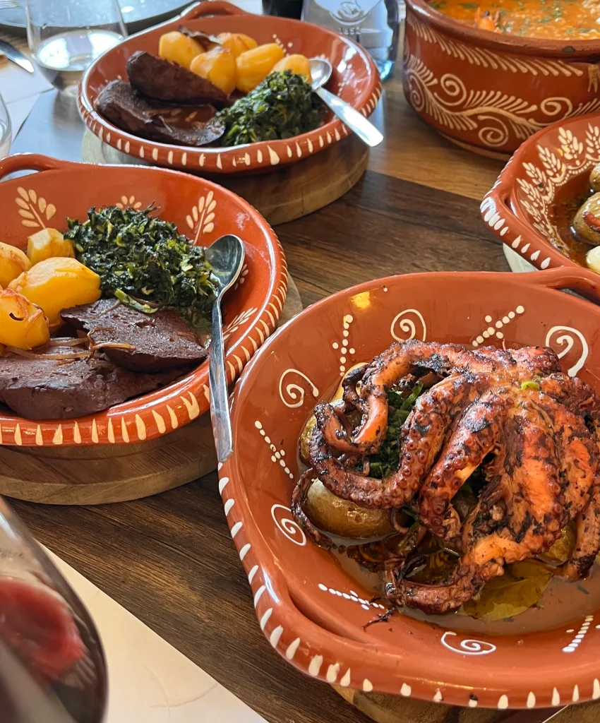 Travessa de barro com polvo assado e batatas a murro ao lado de pratos tradicionais de barro com carne e migas.