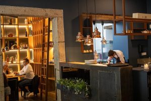 Sala de restaurante com estantes decorativas, mesa com clientes e cozinha aberta com chef a preparar pratos.