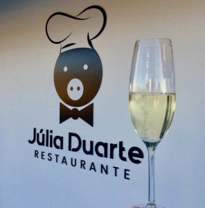 Logótipo do Restaurante Júlia Duarte numa parede, com copo de vinho branco em primeiro plano.