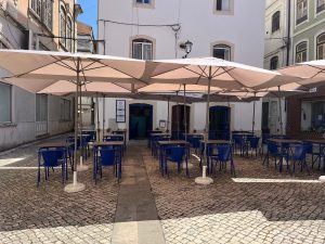 Esplanada com mesas e cadeiras azuis sob guarda-sóis, situada num pátio de calçada junto a edifícios tradicionais.