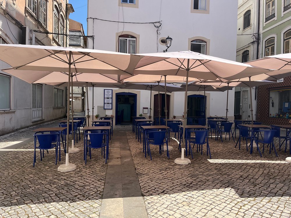 Esplanada com mesas e cadeiras azuis sob guarda-sóis, situada num pátio de calçada junto a edifícios tradicionais.