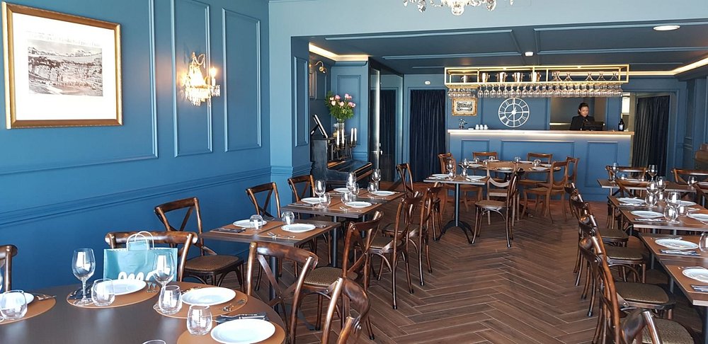 Sala com mesas montadas, chão em padrão espinha, paredes azuis, quadros e bar com garrafeira.